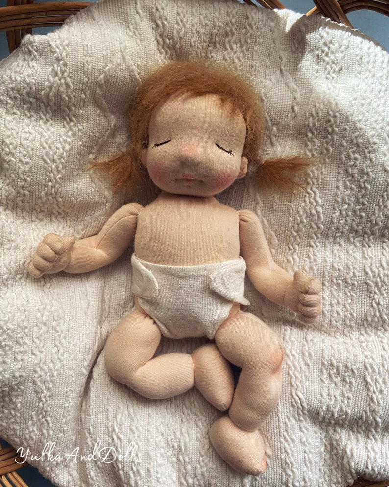 Pode incluir: Uma boneca de pano feita &agrave; m&atilde;o com uma fralda branca, cabelo castanho e olhos fechados, deitada em um cobertor de malha branca.