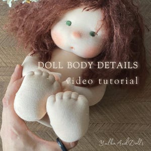 May include: A fabric doll with a detailed face, brown hair, green eyes, and rosy cheeks. The doll's body is made of soft fabric. The text "DOLL BODY DETAILS video tutorial" is visible. The doll is held in a person's hand.
