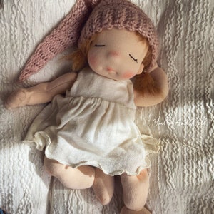 Pode incluir: Uma boneca feita &agrave; m&atilde;o com um chap&eacute;u de malha rosa, um vestido branco e os olhos fechados, deitada em um cobertor de malha branco.