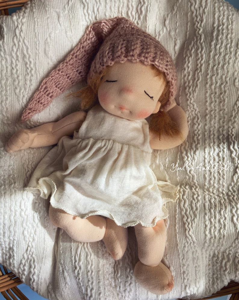 Pode incluir: Uma boneca feita &agrave; m&atilde;o com um chap&eacute;u de malha rosa, um vestido branco e os olhos fechados, deitada em um cobertor de malha branco.