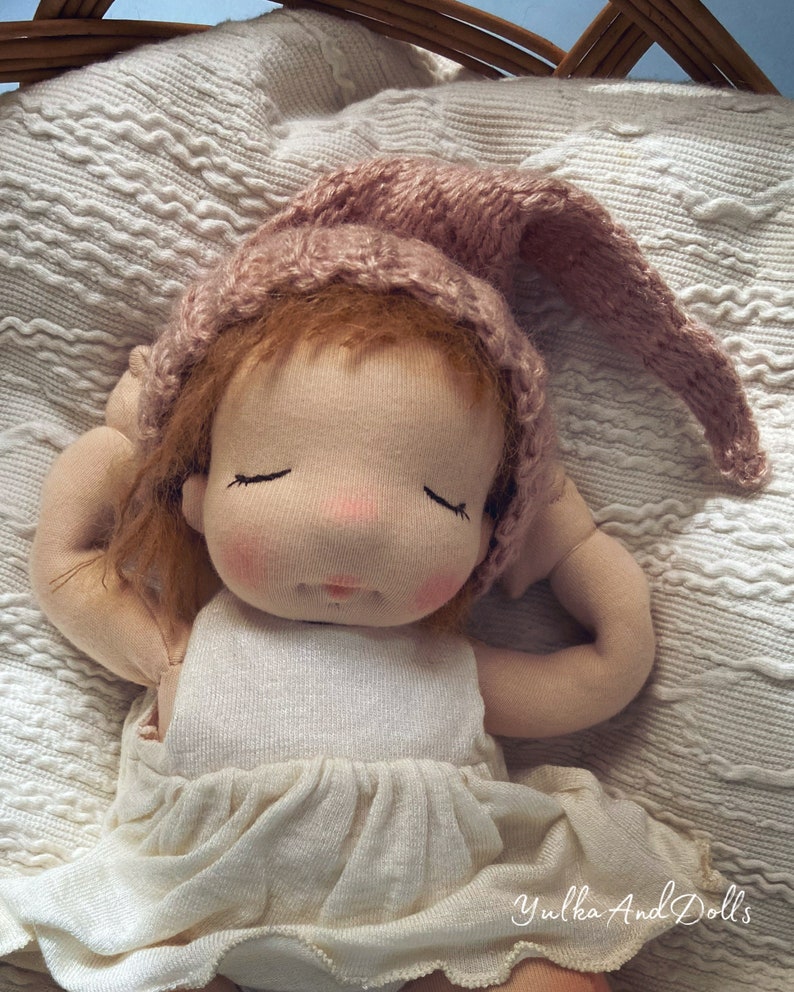 Pode incluir: Uma boneca feita &agrave; m&atilde;o com os olhos fechados, vestindo um chap&eacute;u de malha rosa e um vestido branco, est&aacute; deitada em um cobertor de malha branco. A boneca tem bochechas rosadas e cabelo castanho. O texto "Yulka And Dolls" &eacute; vis&iacute;vel no canto inferior direito da imagem.