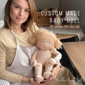 Puede incluir: Una muñeca de bebé hecha a medida, de 50 cm de altura, con cabello rubio peinado en dos moños. La muñeca tiene ojos azules y está vestida con una tela beige claro. El texto "Custom Made Baby Doll" y el nombre de la tienda "Yulka And Dolls" están incluidos.