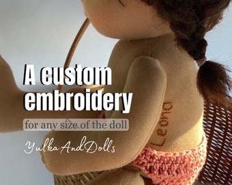 May include: A custom embroidery for any size doll. The doll is sitting in a wicker basket and has a pink crocheted diaper. The embroidery says "Leona".