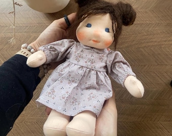 May include: A handmade doll with brown hair styled in two buns. The doll has blue eyes, rosy cheeks, and a small smile. It wears a light purple dress with a floral pattern. The doll's arms and legs are a light beige color.