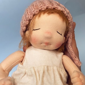 Pode incluir: Uma boneca de pano feita &agrave; m&atilde;o com os olhos fechados, usando um chap&eacute;u de malha rosa e um vestido branco. A boneca est&aacute; sendo segurada por uma m&atilde;o.
