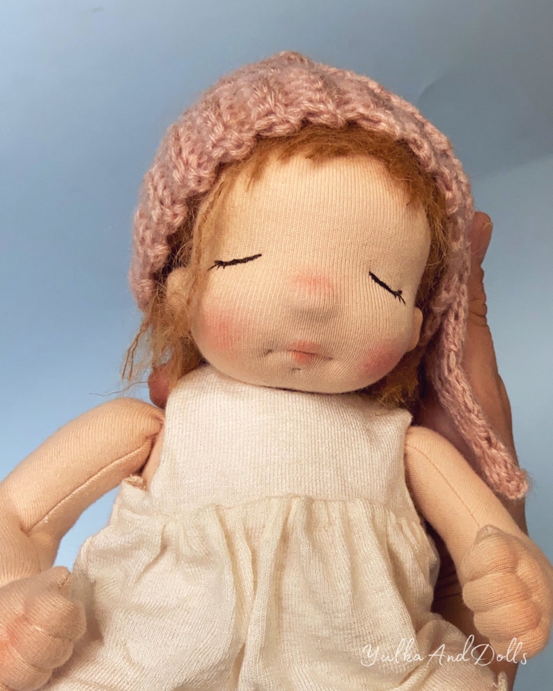 Pode incluir: Uma boneca de pano feita &agrave; m&atilde;o com os olhos fechados, usando um chap&eacute;u de malha rosa e um vestido branco. A boneca est&aacute; sendo segurada por uma m&atilde;o.