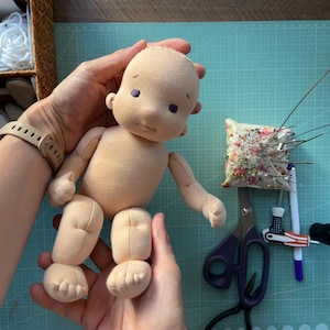 May include: A handmade cloth doll with a peach-colored body and limbs, held in hands. The doll has purple eyes and a simple face. Sewing tools, including scissors and a pin cushion, are on a green cutting mat.