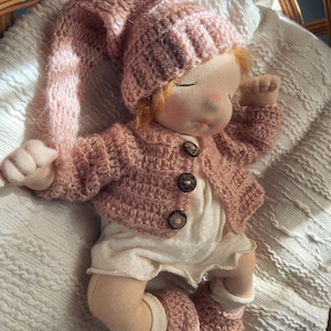 Pode incluir: Uma boneca de pano feita &agrave; m&atilde;o com um chap&eacute;u, um casaco e botinhas de croch&ecirc; rosa. A boneca est&aacute; deitada em um cobertor branco com uma cesta de vime no fundo.
