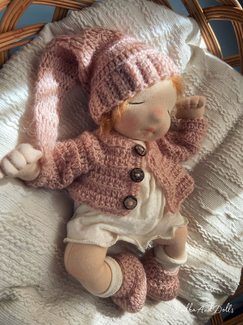 Pode incluir: Uma boneca de pano feita &agrave; m&atilde;o com um chap&eacute;u, um casaco e botinhas de croch&ecirc; rosa. A boneca est&aacute; deitada em um cobertor branco com uma cesta de vime no fundo.