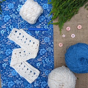 May include: A craft scene featuring a cream-colored crocheted scarf, yarn balls, a blue crochet hook, and pink buttons. The scarf is laid on a blue floral fabric, with a ball of cream yarn and a ball of blue yarn. Green sprigs and buttons add detail.