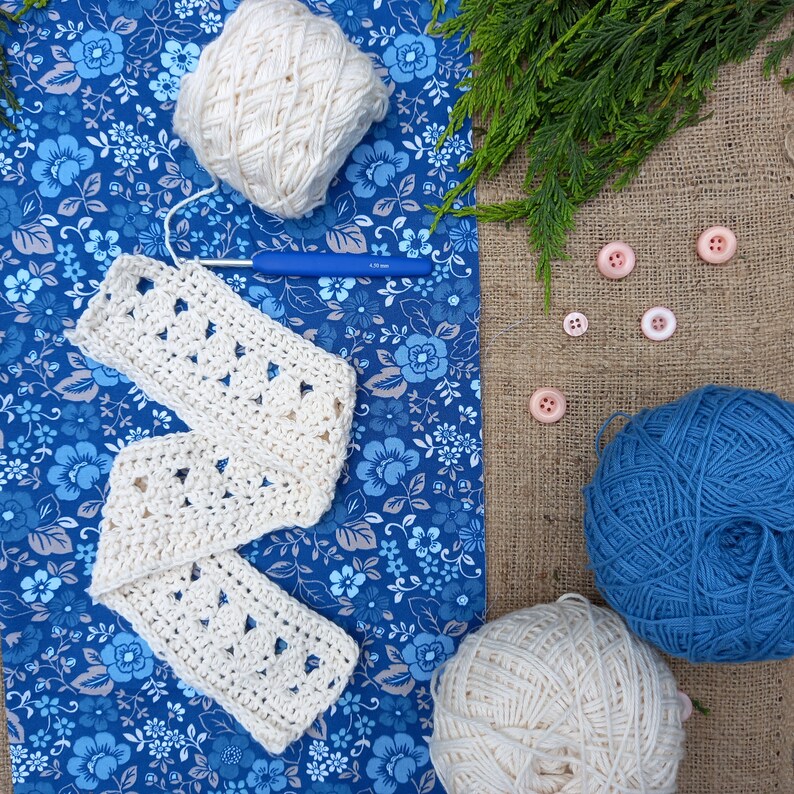 May include: A craft scene featuring a cream-colored crocheted scarf, yarn balls, a blue crochet hook, and pink buttons. The scarf is laid on a blue floral fabric, with a ball of cream yarn and a ball of blue yarn. Green sprigs and buttons add detail.