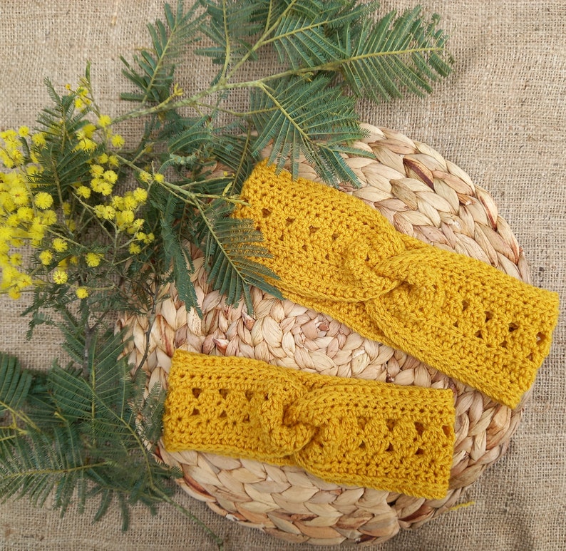 May include: Two mustard yellow crochet headbands with a twisted design, resting on a woven mat. The headbands are surrounded by sprigs of yellow flowers and green leaves, set against a burlap background.