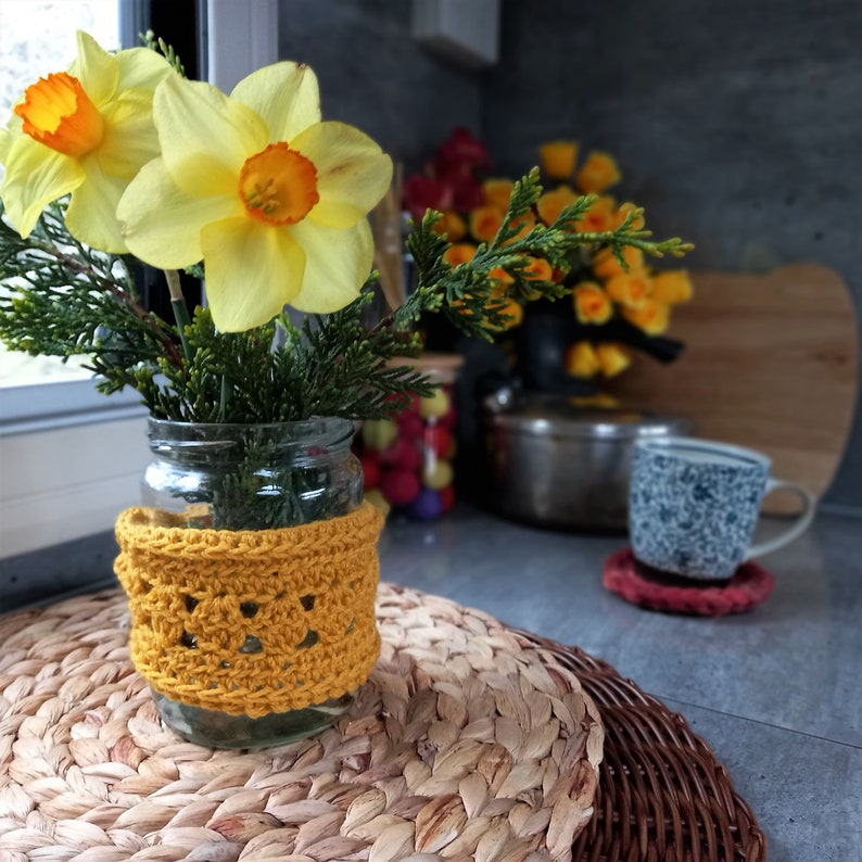 May include: A glass jar vase with yellow daffodils and greenery, adorned with a mustard-colored crocheted cozy. The vase sits on a woven placemat, with a blurred background featuring a mug and other kitchen items.