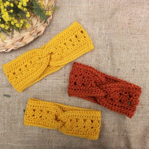 May include: Three handmade crochet headbands in shades of yellow and rust-orange are displayed on a textured surface. The headbands feature a twisted design and openwork detailing. The headbands are a cozy accessory.