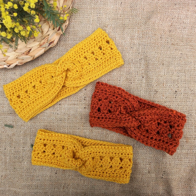 May include: Three handmade crochet headbands in shades of yellow and rust-orange are displayed on a textured surface. The headbands feature a twisted design and openwork detailing. The headbands are a cozy accessory.