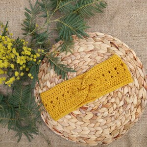 May include: Mustard yellow crochet headband with a twisted design. The headband is made of a soft, textured yarn and features a series of small, openwork details. The headband is displayed on a woven, circular placemat, with sprigs of yellow flowers and green leaves.