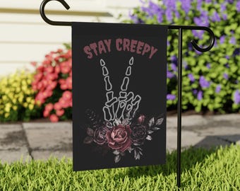 Puede incluir: Bandera de jardín negra con el texto "STAY CREEPY" sobre una mano de esqueleto haciendo el signo de la paz. La bandera también presenta un arreglo floral de rosas y hojas de color rojo oscuro. La bandera se muestra en un soporte de metal negro.