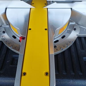 May include: Close-up of a yellow plastic miter saw fence with black screws. The fence is attached to a silver metal base with a circular protractor.