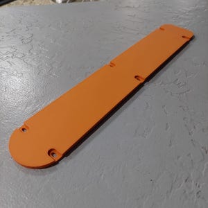 May include: An orange, rectangular object with rounded ends and several holes. The object is lying on a gray surface. The object appears to be a cover or a protective piece.