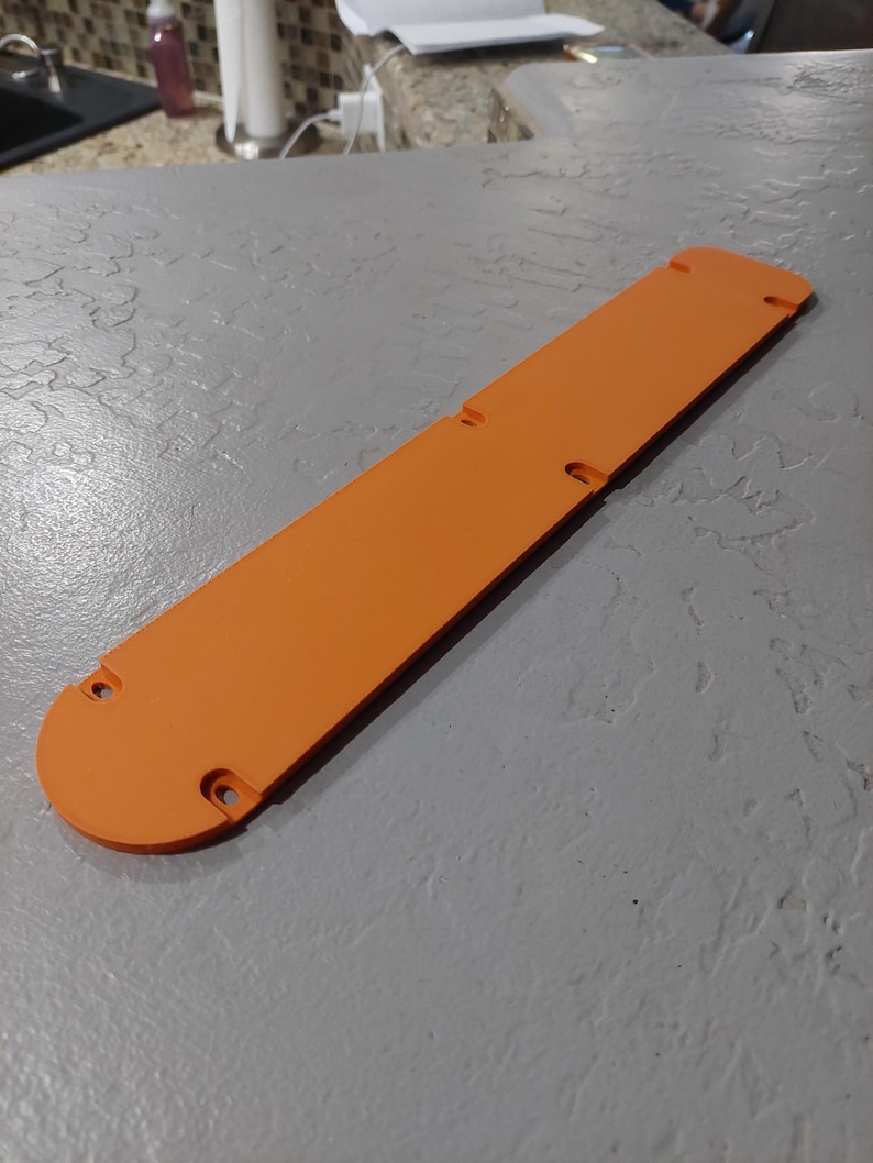 May include: An orange, rectangular object with rounded ends and several holes. The object is lying on a gray surface. The object appears to be a cover or a protective piece.