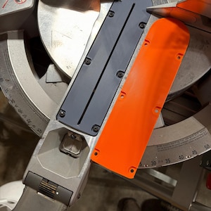 May include: A close-up of a miter saw with a silver blade and a gray and orange plastic guard. The saw has a degree scale and is designed for cutting wood at various angles.
