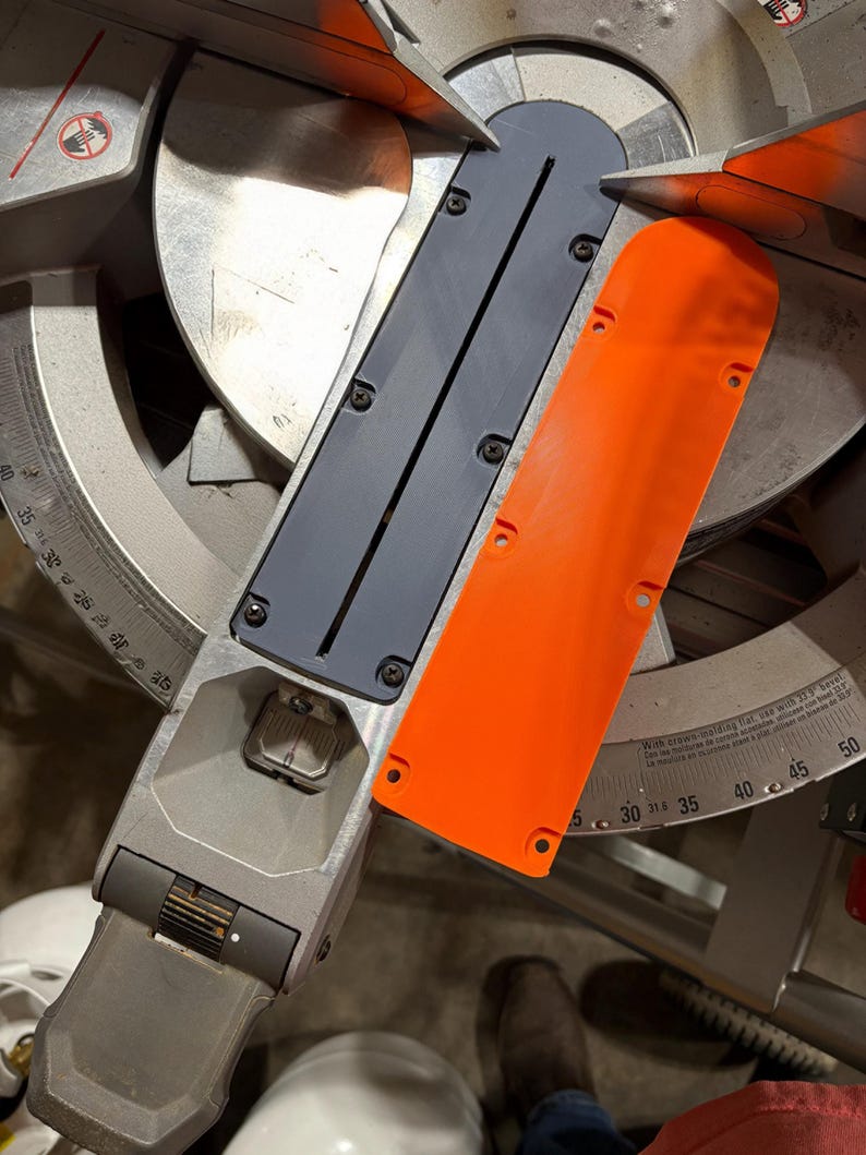 May include: A close-up of a miter saw with a silver blade and a gray and orange plastic guard. The saw has a degree scale and is designed for cutting wood at various angles.