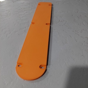 May include: An orange, elongated object with a rounded end and two circular holes. The object has two rectangular cutouts and is lying on a textured, gray surface. The object appears to be made of plastic.