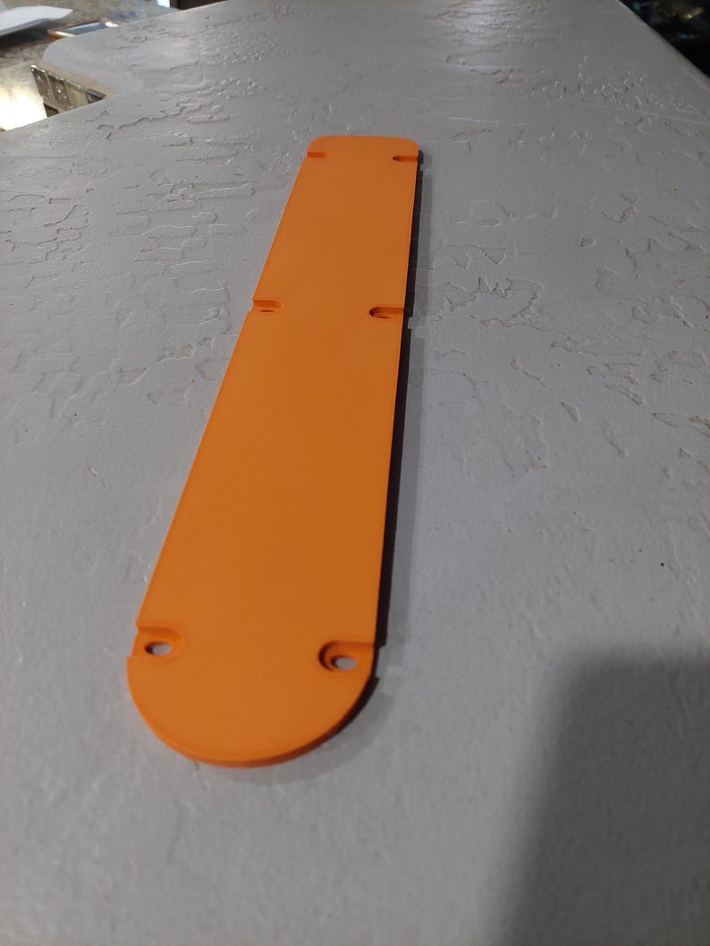 May include: An orange, elongated object with a rounded end and two circular holes. The object has two rectangular cutouts and is lying on a textured, gray surface. The object appears to be made of plastic.