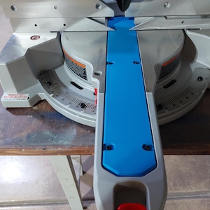 May include: A silver and blue miter saw with a blue center section and a red button. The saw has a circular blade and a degree scale for angled cuts. The saw is on a wooden surface.