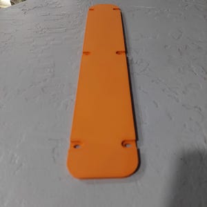 May include: Bright orange, rectangular object with rounded ends and several small holes. The object is long and narrow, with a flat surface. The color is a vibrant, solid orange.