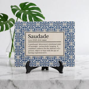 May include: A decorative sign with a blue and white patterned border and the word "Saudade" in the center. The definition of "Saudade" is printed below the word. The sign is displayed on a black easel.