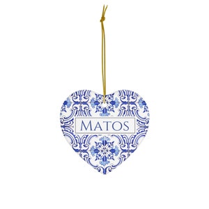 May include: A white heart-shaped ceramic ornament with a blue and white patterned design. The ornament has a gold string for hanging and the name "MATOS" written in blue on the front.