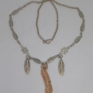 May include: A silver-toned necklace featuring a chain with integrated metal feather charms, decorative filigree links, and a central pendant shaped like a carved light brown feather. The necklace includes a lobster claw clasp closure.