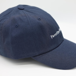 May include: A navy blue baseball cap with a curved peak. The cap has the embroidered text "Favorite Day" in white on the side. It features a button on top and an adjustable strap at the back.