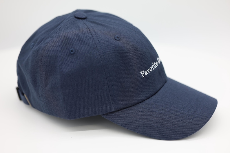 May include: A navy blue baseball cap with a curved peak. The cap has the embroidered text "Favorite Day" in white on the side. It features a button on top and an adjustable strap at the back.
