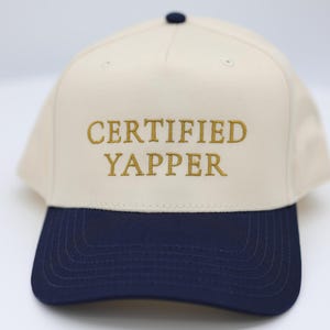 May include: A baseball cap with a cream-coloured crown and a navy blue brim. The phrase "CERTIFIED YAPPER" is embroidered in gold thread across the front. The cap has a navy blue button on top.