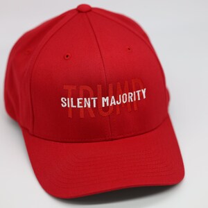 Könnte beinhalten: Rote Baseballkappe mit den Wörtern "TRUMP SILENT MAJORITY" in Weiß gestickt. Die Kappe besteht aus einem einfarbigen roten Stoff und hat einen gebogenen Schirm. Der Text ist mittig auf der Vorderseite der Kappe angebracht.