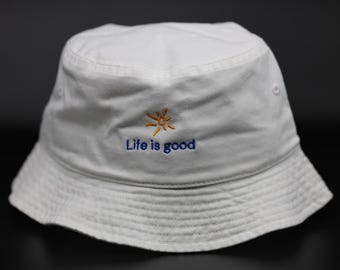 May include: White bucket hat with the embroidered phrase "Life is good" in blue, and a small orange sun graphic. The hat is made of a soft fabric and has a classic, casual style.