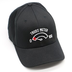May include: Black baseball cap with a white embroidered design of a trust meter with a red needle pointing to zero. The text "TRUST METER" is above the meter and "100" is to the right of the meter.