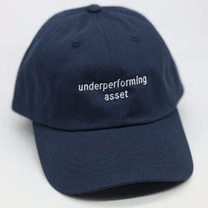 May include: A navy blue baseball cap with the text "underperforming asset" embroidered in white on the front.