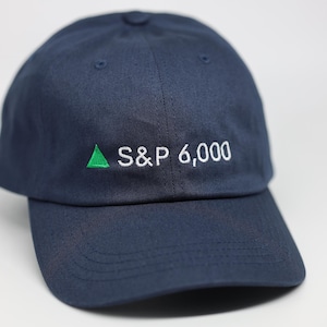 May include: A navy blue baseball cap featuring the embroidered text "S&P 6,000" in white, with a green upward-pointing triangle. The cap has a curved peak and a button on top.