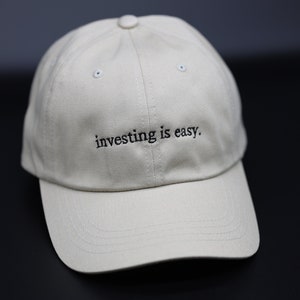 May include: A beige baseball cap with the embroidered text "investing is easy." The cap has a curved peak and a button on top. The background is a dark grey.