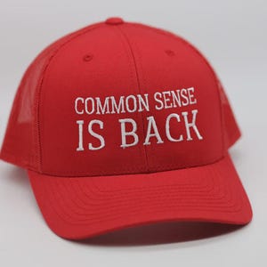 Może przedstawiać: Czerwona czapka trucker z siateczką z tyłu. Na czapce znajduje się białe haftowane zdanie "COMMON SENSE IS BACK".