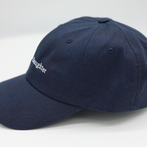 May include: A navy blue baseball cap with a curved peak. The word "daughter" is embroidered in white on the front. The hat has a classic six-panel design with a button on top.