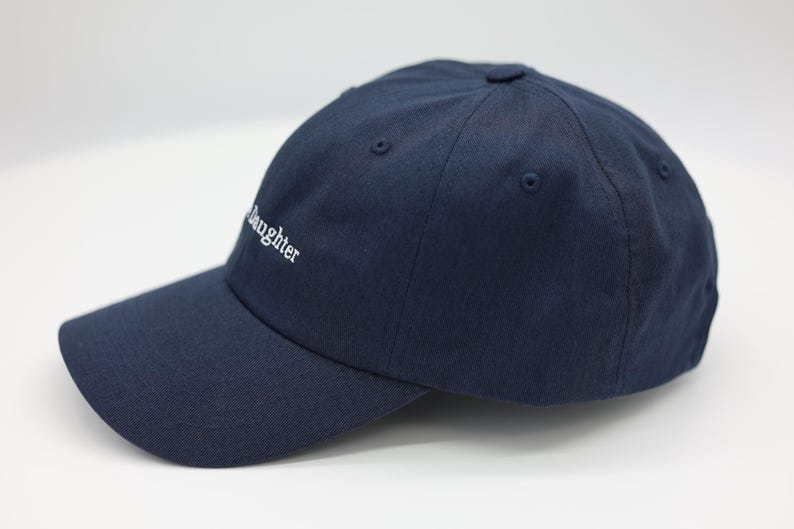 May include: A navy blue baseball cap with a curved peak. The word "daughter" is embroidered in white on the front. The hat has a classic six-panel design with a button on top.