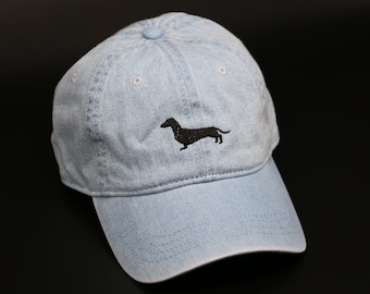 Puede incluir: Una gorra de béisbol de mezclilla azul claro con una silueta de dachshund bordada en negro.