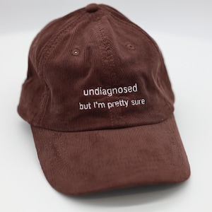 May include: A brown corduroy baseball cap with a curved brim. The cap features the embroidered text "undiagnosed but I'm pretty sure" in white. The cap is a solid brown color.