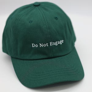 以下が含まれることがあります： 白い文字で「Do Not Engage」と刺繍された濃い緑色の野球帽。帽子はカーブしたつばと、布で覆われたボタンが付いています。帽子は丈夫な生地で作られています。