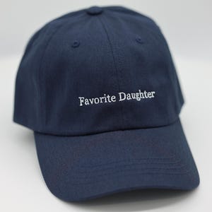 May include: A navy blue baseball cap with the embroidered text "Favorite Daughter" in white. The cap features a curved peak and a button on top. A casual accessory.