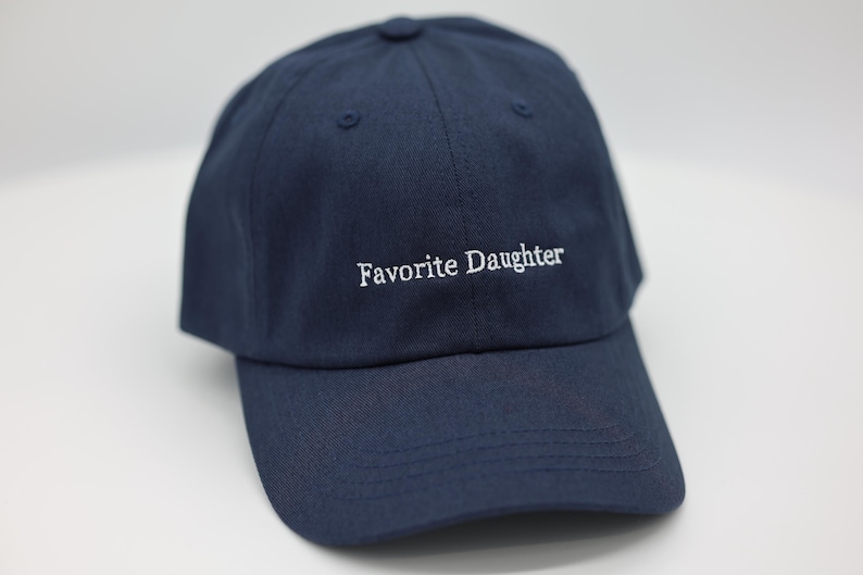 May include: A navy blue baseball cap with the embroidered text "Favorite Daughter" in white. The cap features a curved peak and a button on top. A casual accessory.